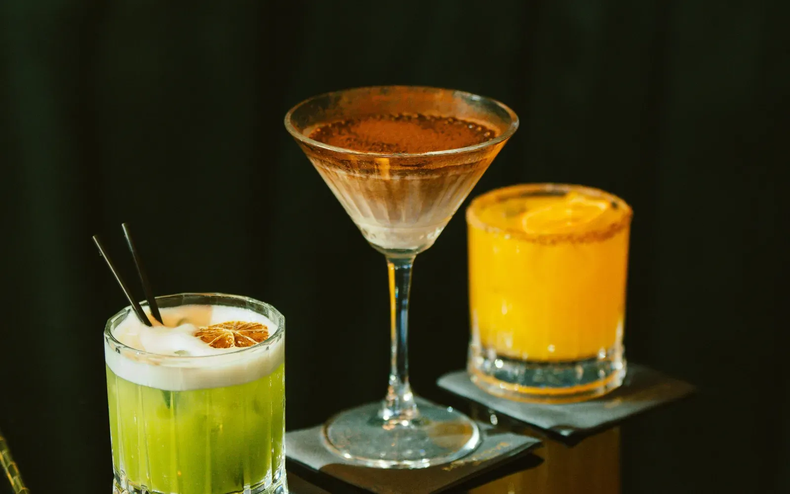 Drei bunte Cocktails auf einer dunklen, spiegelnden Bar – Swingerclub Preise und was im Eintritt steckt